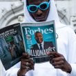 A man in a white hoodie and blue sunglasses reads a magazine about the richest people in the world.