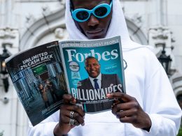 A man in a white hoodie and blue sunglasses reads a magazine about the richest people in the world.