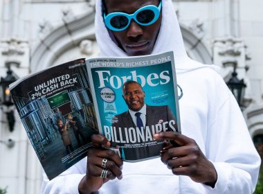 A man in a white hoodie and blue sunglasses reads a magazine about the richest people in the world.