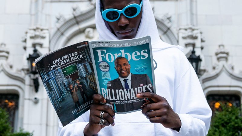 A man in a white hoodie and blue sunglasses reads a magazine about the richest people in the world.