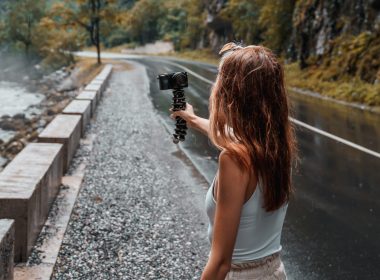 Woman vlogger recording herself next to a wooded mountainside, road, and stream.