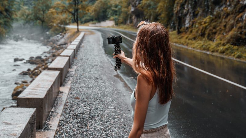 Woman vlogger recording herself next to a wooded mountainside, road, and stream.