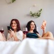 Two women enjoying red wind and relaxing together in a big bed with white sheets