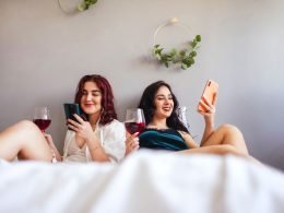 Two women enjoying red wind and relaxing together in a big bed with white sheets