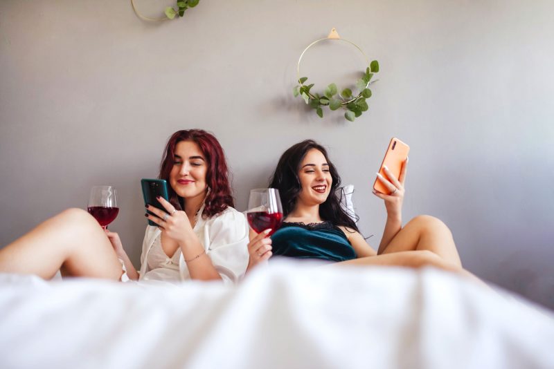 Two women enjoying red wind and relaxing together in a big bed with white sheets