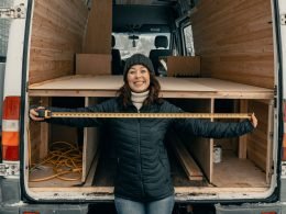 Woman holding a measuring tape while standing in front of an RV in construction in cool weather.