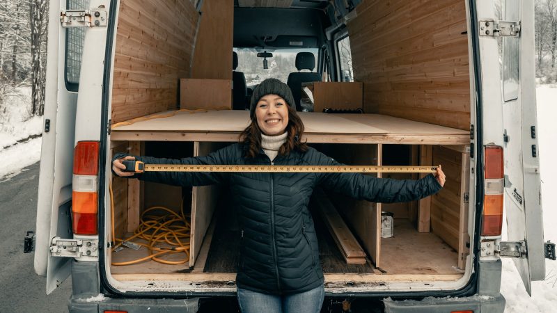 Woman holding a measuring tape while standing in front of an RV in construction in cool weather.