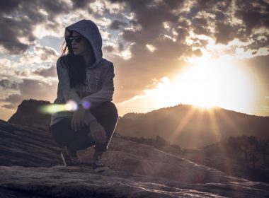Hooded woman kneeling in the desert looking around as the sun starts to set.