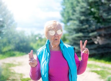 Hip, mature woman with white hair, aviators and a bight blue vest walking in a trail in the woods giving peace signs with her hands.