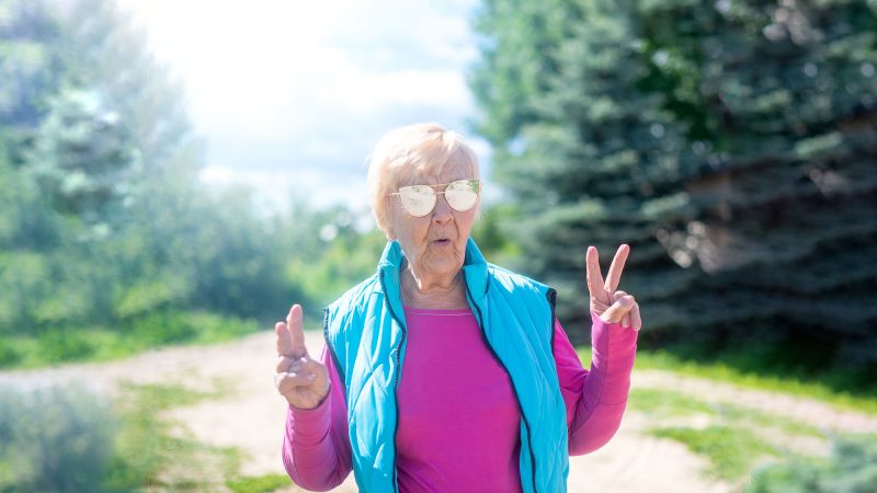 Hip, mature woman with white hair, aviators and a bight blue vest walking in a trail in the woods giving peace signs with her hands.