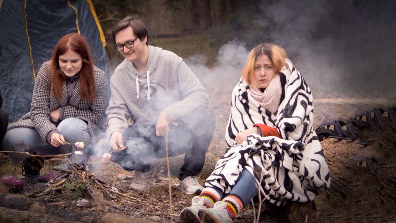 A group of friends tries to start a fire while camping and one of them is miserable wrapped in blankets with smoke blowing in her face.