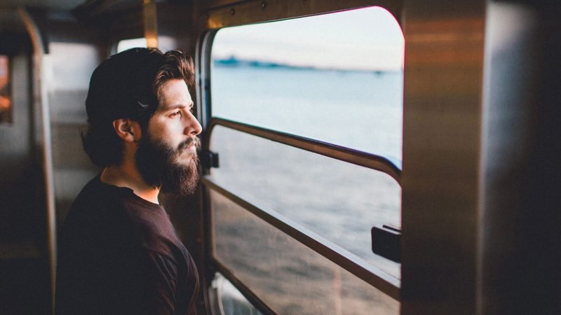 A younbg man with a full head of hair and beard stares out the window of an RV over lake.