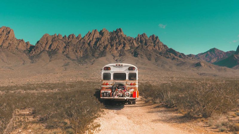 The back of a white skoolie in the desert with rocky mountains in the distance and kids' bikes on mounted on the back.