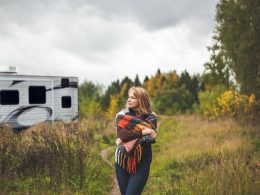 An adult woman wraps a plaid blanket over her shoulders as she walks in contemplation through the woods away fro her travel trailer.