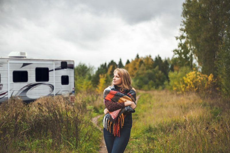 An adult woman wraps a plaid blanket over her shoulders as she walks in contemplation through the woods away fro her travel trailer.