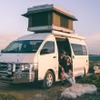 A couple sits in the open door of their camper van with a pop up top.