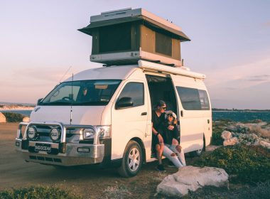 A couple sits in the open door of their camper van with a pop up top.