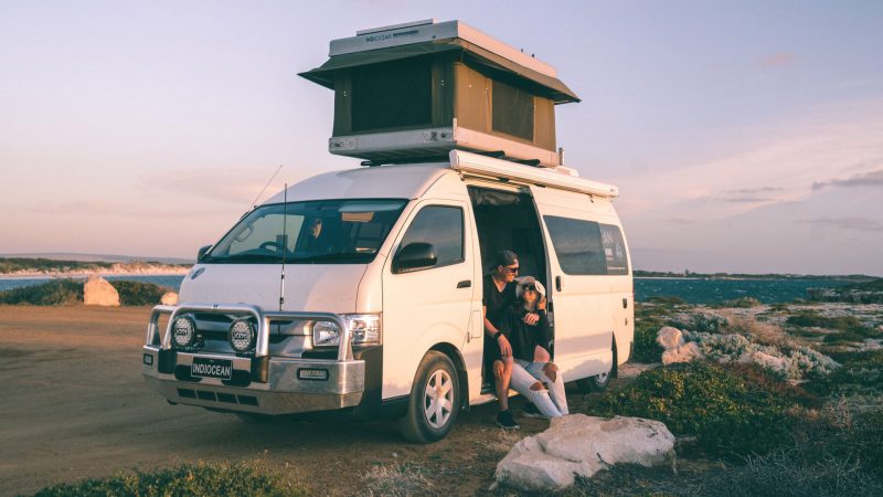 A couple sits in the open door of their camper van with a pop up top.