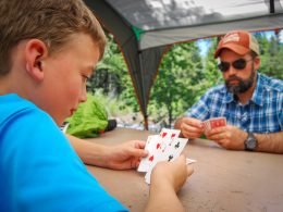 A father and son play a card game at a picnic table while on a camping trip.