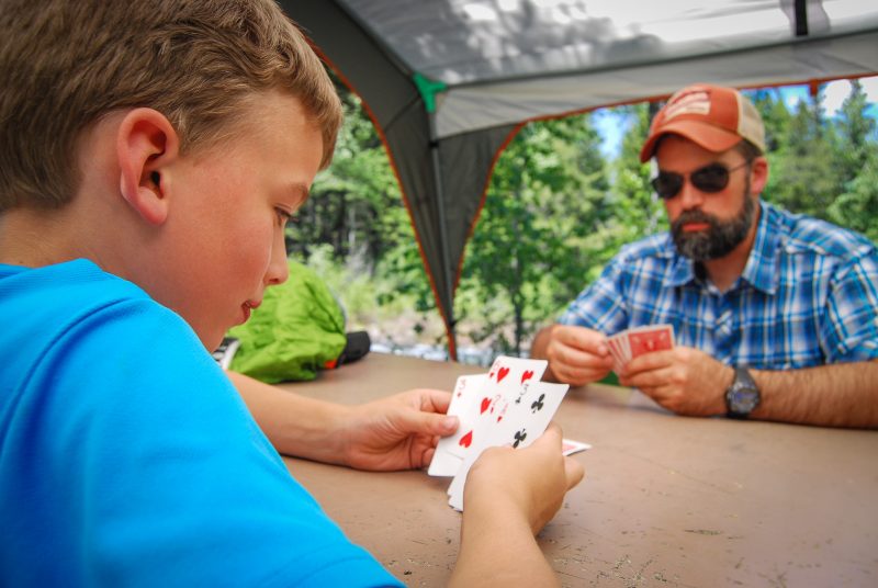 A father and son play a card game at a picnic table while on a camping trip.