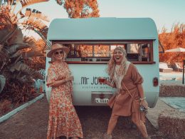 Two women dressed in boho garb outside their vintage mobile with cocktails.