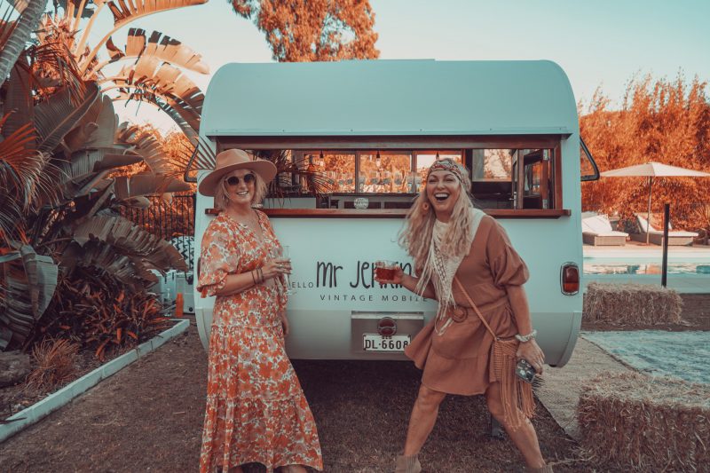 Two women dressed in boho garb outside their vintage mobile with cocktails.