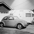 A black and white old photo of a 1960s VW bug and a travel trailed parked in a driveway in California