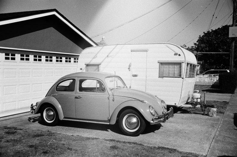 A black and white old photo of a 1960s VW bug and a travel trailed parked in a driveway in California