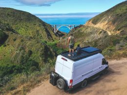 Two people stand atop their van overlooking a beautiful landscape with a valley and bridge in the distance. They are living the #vanlife