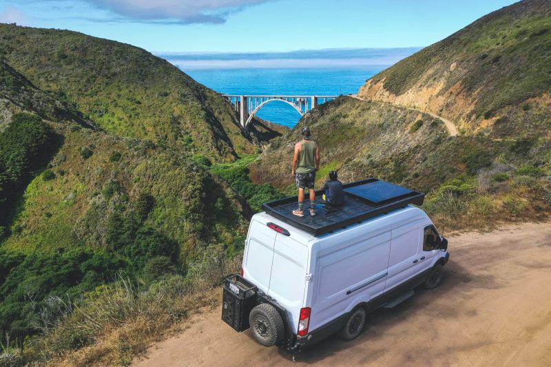Two people stand atop their van overlooking a beautiful landscape with a valley and bridge in the distance. They are living the #vanlife