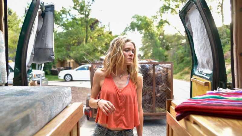 A tired woman looks in from the back doors of the van she's living in.