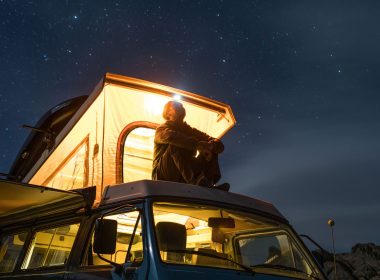 A man sits on the top of his camper van glowing with a warm light in the against the night sky in Alabama Hills.