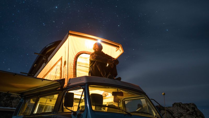 A man sits on the top of his camper van glowing with a warm light in the against the night sky in Alabama Hills.