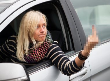 Angry woman aggressively gives the middle finer out her car window as she drives.