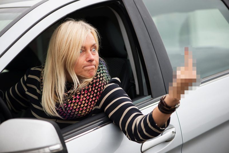 Angry woman aggressively gives the middle finer out her car window as she drives.