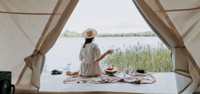 A woman in white linen and a hat enjoys wine and h'ordeuvres, she is glamping in a tent on a lake.