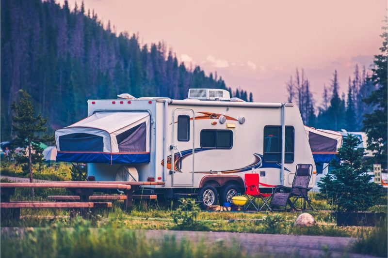 A hybrid camper parked in a campground near forested mountains at dusk.
