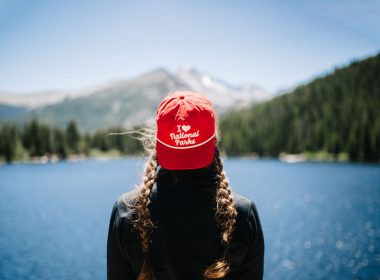 A woman wears a red 'I love national parks' ball cap backwards as she overlooks a sparkling lake, vast forests, and towering mountains in the distance.