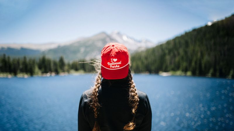 A woman wears a red 'I love national parks' ball cap backwards as she overlooks a sparkling lake, vast forests, and towering mountains in the distance.