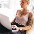 Young, beautiful, tattooed woman sits corss-legged with her laptop