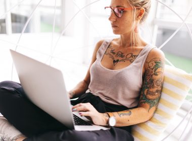 Young, beautiful, tattooed woman sits corss-legged with her laptop