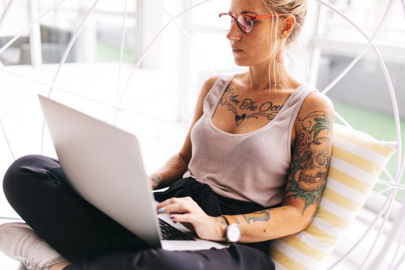 Young, beautiful, tattooed woman sits corss-legged with her laptop