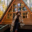 A young, boho woman wrapped in a blanket sits outside of her a-frame tiny home.