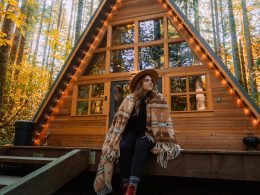 A young, boho woman wrapped in a blanket sits outside of her a-frame tiny home.