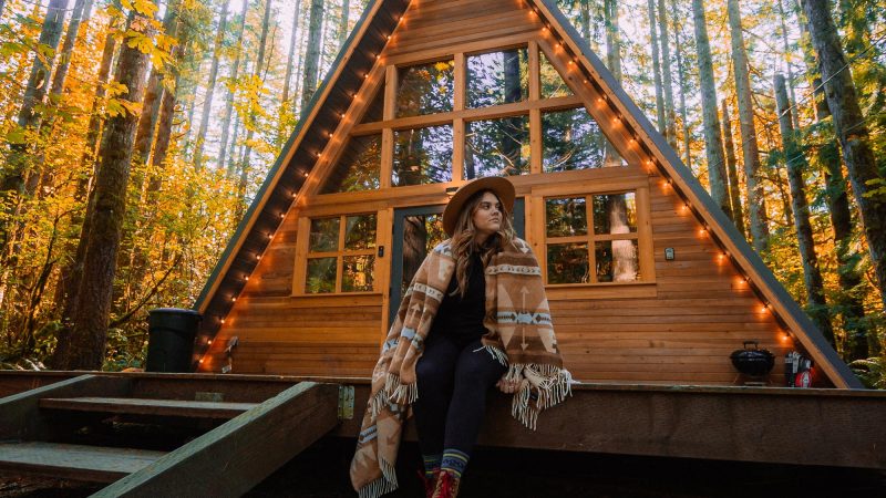 A young, boho woman wrapped in a blanket sits outside of her a-frame tiny home.