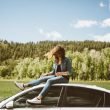 A woman sits on top of her car with forested hills behind her as the wind blows her hair freely into her face.