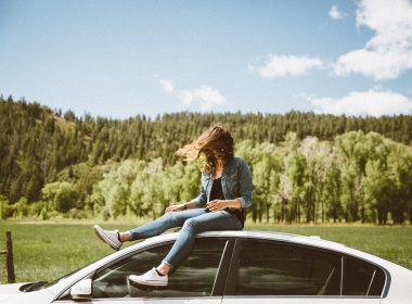 A woman sits on top of her car with forested hills behind her as the wind blows her hair freely into her face.