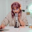 Woman with pink hair and a sweater recording a podcast at a desk.