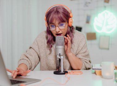 Woman with pink hair and a sweater recording a podcast at a desk.