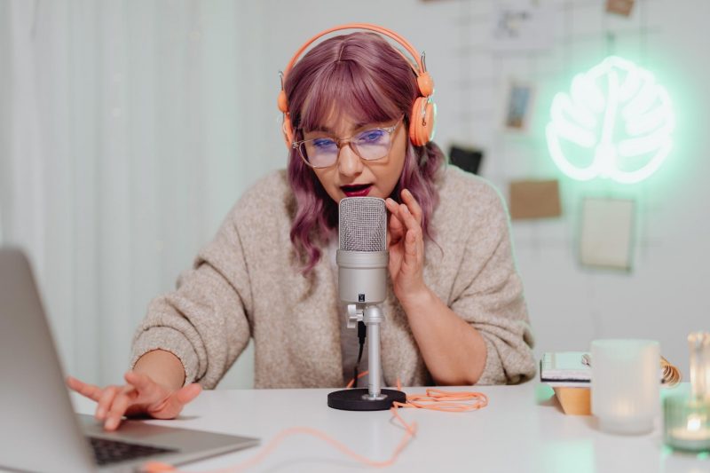 Woman with pink hair and a sweater recording a podcast at a desk.